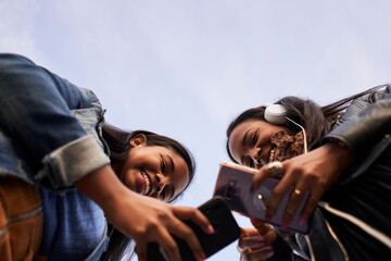 Low angle of two young African American women with headphones listening to music with mobil outdoors. Isolated silhouette in the sky. Copy space