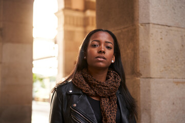 Portrait of young beautiful African American woman looking at camera with serious face outdoors with the sun behind her. Serious millennial girl standing on the city street. People and emotions