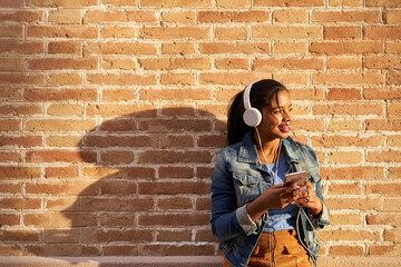 Portrait of young African American woman with headphones listening to music from her mobile phone in front of a brick wall.
