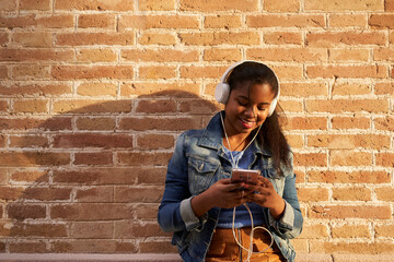 Portrait of young African American woman with headphones listening to music while using her mobile phone in front of a brick wall.
