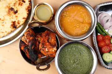Spicy red Chicken grilled tikka tandoori nan bread green spinach curry yogurt sauce dal tomato cucumber onion salad set in metal stainless steel brass copper pot on light wood background