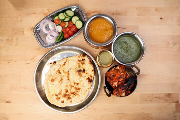 Spicy red Chicken grilled tikka tandoori nan bread green spinach curry yogurt sauce dal tomato cucumber onion salad set in metal stainless steel brass copper pot on light wood background