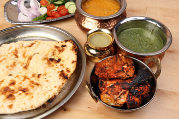 Spicy red Chicken grilled tikka tandoori nan bread green spinach curry yogurt sauce dal tomato cucumber onion salad set in metal stainless steel brass copper pot on light wood background