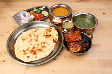 Spicy red Chicken grilled tikka tandoori nan bread green spinach curry yogurt sauce dal tomato cucumber onion salad set in metal stainless steel brass copper pot on light wood background