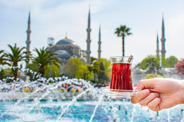 Woman's hand holding cup with Traditional turkish tea in front of Blue mosque (aka Sultanahmet...