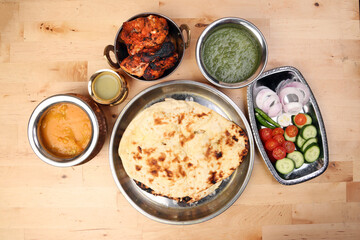 Spicy red Chicken grilled tikka tandoori nan bread green spinach curry yogurt sauce dal tomato cucumber onion salad set in metal stainless steel brass copper pot on light wood background