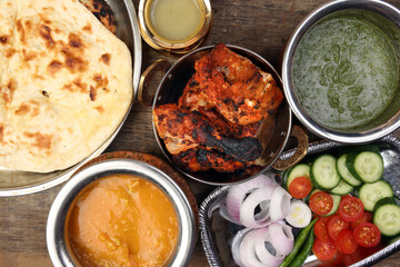 Spicy red Chicken grilled tikka tandoori nan bread green spinach curry yogurt sauce dal tomato cucumber onion salad set in metal stainless steel brass copper pot on rustic vintage wood background