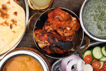 Spicy red Chicken grilled tikka tandoori nan bread green spinach curry yogurt sauce dal tomato cucumber onion salad set in metal stainless steel brass copper pot on rustic vintage wood background