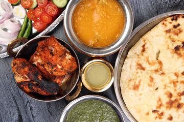 Spicy red Chicken grilled tikka tandoori nan bread green spinach curry yogurt sauce dal tomato cucumber onion salad set in metal stainless steel brass copper pot on black char wood background