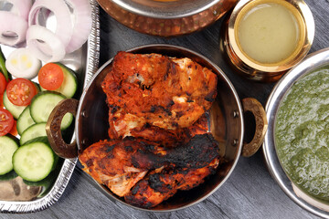 Spicy red Chicken grilled tikka tandoori nan bread green spinach curry yogurt sauce dal tomato cucumber onion salad set in metal stainless steel brass copper pot on black char wood background
