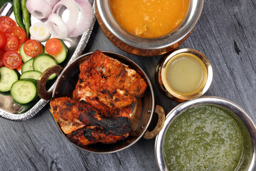 Spicy red Chicken grilled tikka tandoori nan bread green spinach curry yogurt sauce dal tomato cucumber onion salad set in metal stainless steel brass copper pot on black char wood background