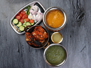Spicy red Chicken grilled tikka tandoori nan bread green spinach curry yogurt sauce dal tomato cucumber onion salad set in metal stainless steel brass copper pot on black char wood background