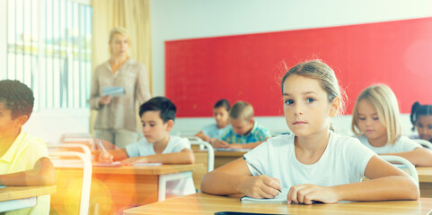 Portrait of focused tween schoolgirl sitting on lesson in classroom, looking confidently at camera