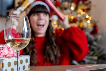 Young girl in Christmas hat and red sweater making a video call to her family to show them Christmas gifts while having a glass of wine with the Christmas tree behind