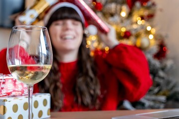 Young girl in Christmas hat and red sweater making a video call to her family to show them Christmas gifts while having a glass of wine with the Christmas tree behind