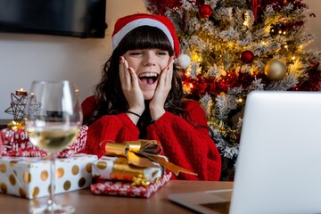 Young girl in Christmas hat and red sweater making a video call to her family to show them Christmas gifts while having a glass of wine with the Christmas tree behind