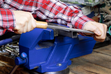 A handyman with file for metal. A locksmith vise. Blue vise on a wooden table. Bench tools. Vice. Men's hands hold a file