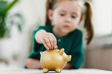 Close up view on child hand, she putting a coin in gold color piggy bank. Selective soft focus