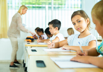 Portrait of positive small female pupil sitting at desk studying in classroom
