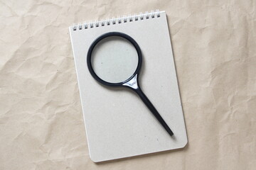 Gray notepad with white coiled spring and magnifier on a background of beige crumpled craft paper. With empty space for text and design