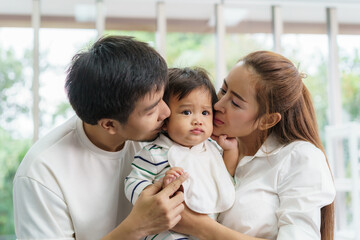 Asian family happy kiss little baby boy in living room at home, young parent and children enjoy love hug standing together..