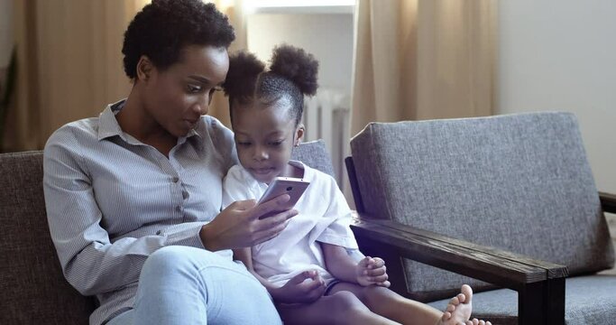 Mixed race mom with African black cute daughter sitting on couch with mobile device smartphone looking at camera screen watching video online on Internet. Woman teaches child to use phone at home