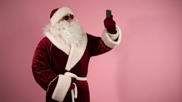 Funny Santa Claus dancing on isolated pink background, man doing online video call on smartphone, talking with online audience and sending christmas wishes. Santa Claus using mobile phone for online
