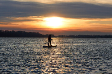 Silhouette of a boy rowing on a SUP (on stand up paddleboard) at sunset in a winter river