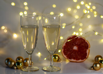two glasses of champagne, pink grapefruit, golden glass balls and various Christmas decorations, illuminated background of a chain of white lights, Christmas waiting time