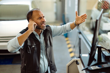 Upset mechanic talking on the phone while reading problematic e-mail on a computer in workshop.