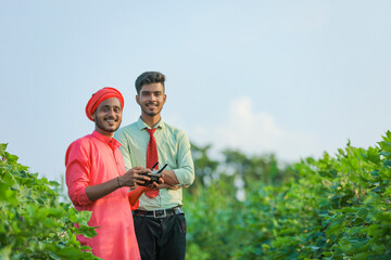 Young indian farmer holding remote in hand and control flying drone with agronomist at agriculture field