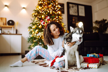 Beautiful woman playing and having fun with her dog while sitting near the christmas tree. Young lady hugging her dog with red christmas hat. Winter holidays. Light around.