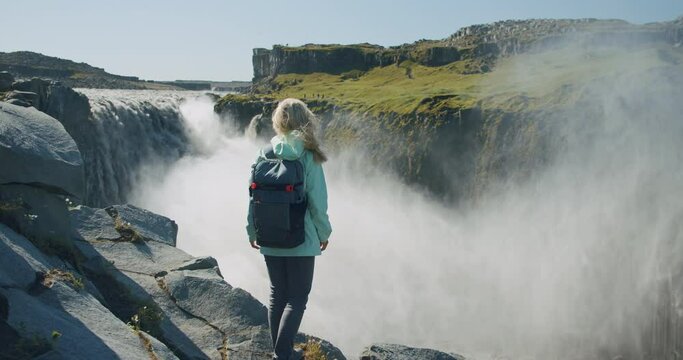 Woman standing at cliff edge looking at powerful Detifoss waterfall in Iceland