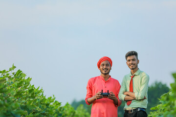 Young indian farmer holding remote in hand and control flying drone with agronomist at agriculture field