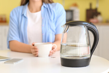 Electric kettle and woman with cup in kitchen, closeup