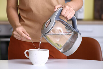 Woman pouring hot boiled water from electric kettle into cup at home