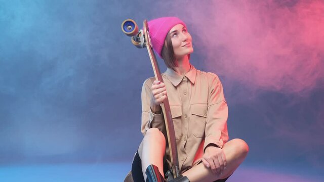 Beautiful young female hipster in stylish clothing and pink hat poses sitting on floor and holding skateboard in colourful and atmospheric background with smoke.