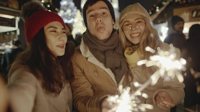 Happy women and man having fun with bengal lights outdoors at the Xmas Fair. Group of friends taking a selfie with smartphone on the Christmas Market. Concept of winter holidays and friendship.