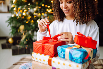 Fototapeta na wymiar Christmas or New Year gift box in the female hands. Happy woman opens presents near the Christmas tree. Winter holidays.