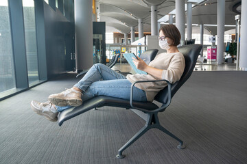 Young female freelancer wearing protective medical mask sits in comfortable chair in airport lounge and works using digital tablet, ready to travel. Life and travel during pandemic.