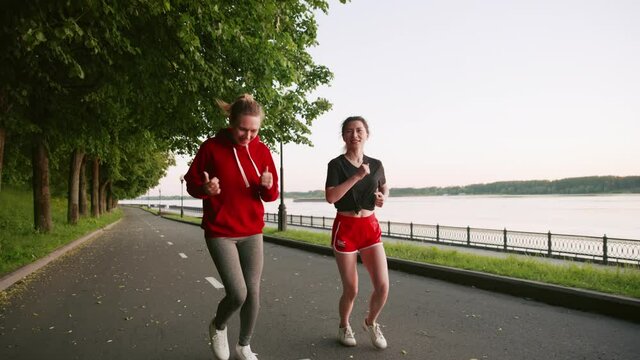 Two young girls in red clothes running in the park. Spend time with friends. Two pretty women communicate with each other while doing sports. Morning or evening jogging along the river embankment.