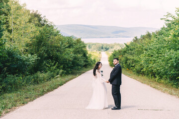 Newlyweds groom in a stylish black suit, the bride in a powdery dress, a photo session in nature, the road against the background of mountains and a reservoir in summer on a sunny day