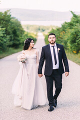 Newlyweds groom in a stylish black suit, the bride in a powdery dress, a photo session in nature, the road against the background of mountains and a reservoir in summer on a sunny day