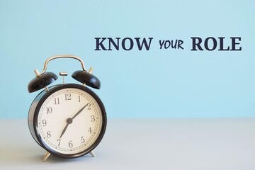 Writing note showing Know Your Role. Business photo showcasing end acting outside who you actually are Play your position. Alarm clock beside a Paper sheet placed on blue backdrop.