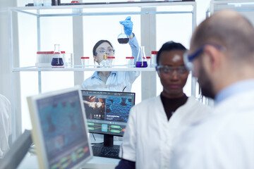 Scientist picks up glass flask with colored liquid in laboratory. Multiethnic team of medical researchers working together in sterile lab wearing protection glasses and gloves.