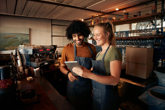 Smiling waiter and waitress wearing apron using digital tablet standing behind counter in cafe