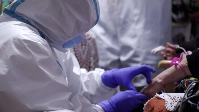 Emergency nurse or paramedic wearing personal protective equipment ppe, nitrile gloves and face mask pricks a patient's hand with a needle to draw blood during coronavirus covid-19 pandemic 