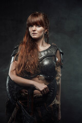 Fototapeta na wymiar Atmospheric portrait of scandinavian amazon with brown hairs dressed in light armour holding two handed axe in dark background.
