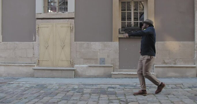 Good looking afro american man in hat dancing while walking along old city street. Bearded happy guy wearing fashionable clothes singing and moving in rhytm . Concept of emotions