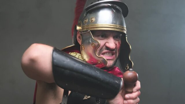 Dressed in military roman clothing bearded imperial soldier pulls out his sword from its scabbard and points it to enemy screaming in dark background.